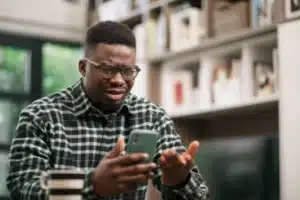 A man indoors visibly annoyed while checking his phone with no signal bars, highlighting mobile reception issues at home.