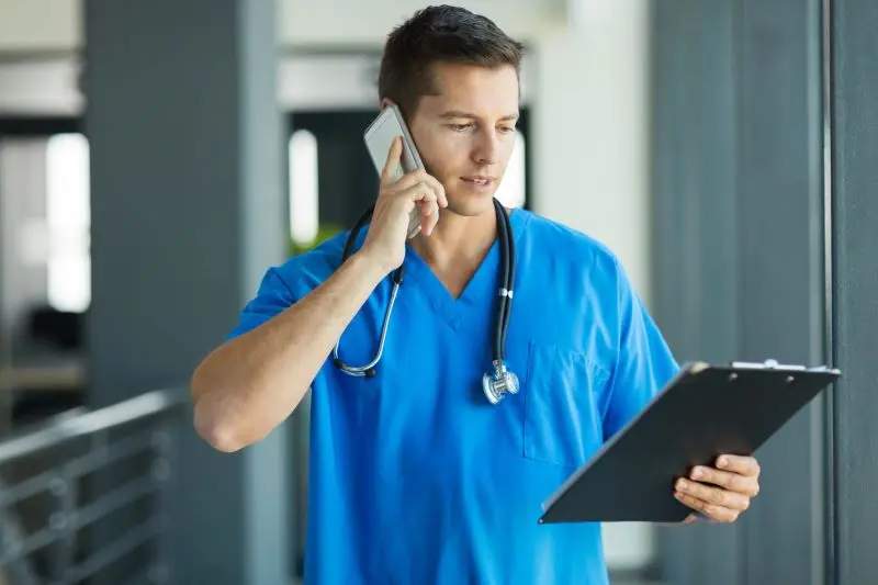 A healthcare professional in blue scrubs talks on the phone and reviews a clipboard in a hallway, efficiently utilizing signal boosters to enhance connectivity for remote healthcare consultations.
